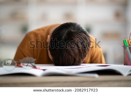Sleeping schooler chubby asian boy laying on desk full of exercise books and stationery, feeling exhausted and bored while studying from home during coronavirus pandemic, copy space