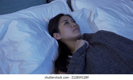 Sleepless Asian Girl Resting In Her Dark Bedroom Is Pulling Up The Blanket And Staring Upward In Contemplation.