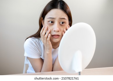 Sleepless Asian woman looking her face in the mirror and worry about dark circles under eyes - Shutterstock ID 2107821761