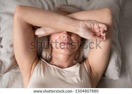 Sleepless mature woman suffering from insomnia close up, lying in bed, older female covering eyes with hands, trying to sleep, nightmares or depression, feeling headache or migraine