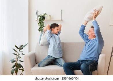 Sleepover With Grandparent. Happy Boy And His Grandfather Fighting With Pillows Having Fun Together Sitting On Couch At Home