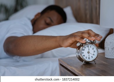 Sleepy African Man Awakening Early In The Morning Turning Off Alarm Clock Lying In Bed At Home, Focus On Alarm-Clock. Black Guy Waking Up In Bedroom Concept. Shallow Depth, Low Light