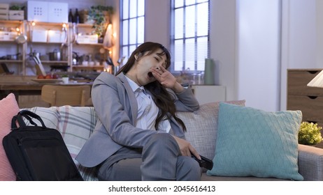 Sleepy Asian Businesswoman With Remote Control In Hand Is Yawning And Propping Head While Watching Tv On Sofa. Genuine Lifestyle.