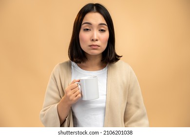 Sleepy asian ethnic female with sleepy eyes holding mug while drinking coffee early in morning against beige background 