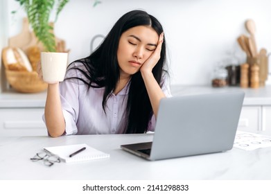 A sleepy Asian girl, freelancer or a student, sits at home in the kitchen at a table with her eyes closed in front of a laptop, holds a cup of coffee in her hand, cannot wake up and start work