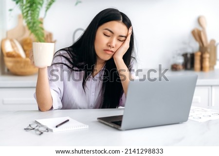 A sleepy Asian girl, freelancer or a student, sits at home in the kitchen at a table with her eyes closed in front of a laptop, holds a cup of coffee in her hand, cannot wake up and start work Foto stock © 
