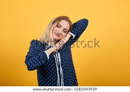 Sleepy beauty caucasian young lady holding hand on head, doing sleeping gesture over isolated orange background in blue shirt with white polka dot. Lifestyle concept