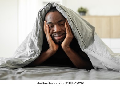 Sleepy Black Guy Covered With Blanket Lying Suffering From Sleeplessness Touching Face In Bed In Modern Bedroom At Home. Insomnia And Sleeping Routine Issue Concept