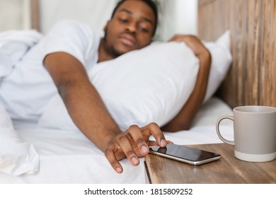 Sleepy Black Man Reaching For Smartphone Turning Off Alarm Clock Waking Up Early Lying In Bed In Bedroom At Home In The Morning. Lack Of Sleep And Rest Concept. Selective Focus, Shallow Depth