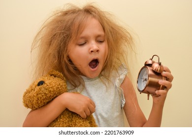 A sleepy child yawns with an alarm clock in his hands. Early rise to school or kindergarten. The kid was awakened early in the morning with tousled hair. The little girl did not get enough sleep. 