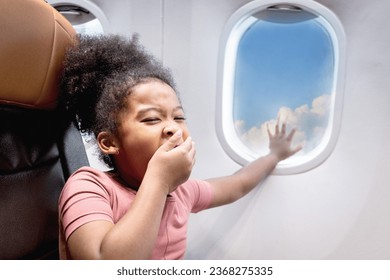 Sleepy curly hair African girl child passenger sitting in comfortable seat next to window inside airplane, kid yawning and reaching hand out to touch window glass, feeling sleepy and needing rest - Powered by Shutterstock