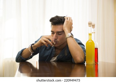 Sleepy, drunk young man sitting drinking alone at a table with two bottles of liquor alongside him sipping from shot glass to drown his sorrows