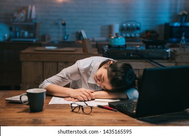 Sleepy exhausted asian woman employee working at wooden kitchen desk with laptop computer. lady eyes are closing and about to fall asleep. sleep deprivation and overtime working concept in midnight.
