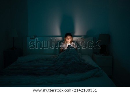 Sleepy exhausted woman lying in bed using smartphone, can not sleep. Insomnia, addiction concept. Sad girl bored in bed scrolling through social networks on mobile phone late at night in dark bedroom.