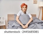 sleepy extravagant queer person in homewear with red hair sitting on bed and looking at camera