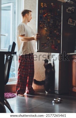 Sleepy full length young guy open fridge looking for breakfast food drinking coffee at early morning in kitchen. Millennial man feed cute funny black cat at cozy home modern bright apartment.