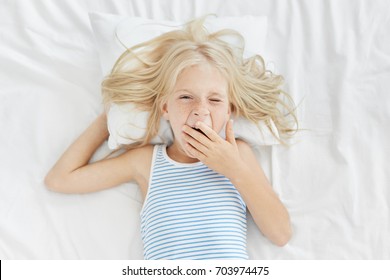 Sleepy girl waking up early in morning, covering mouth with hand while yawning, going to school or kindergarten. Blonde charming child in sailor T-shirt lying on white bed clothes, just awaking - Powered by Shutterstock