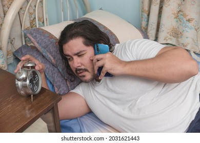 A Sleepy And Groggy Man Lying In Bed Checks The Time On His Alarm Clock While Talking To A Client. Freelancer Taking Up Rush Work In The Morning.