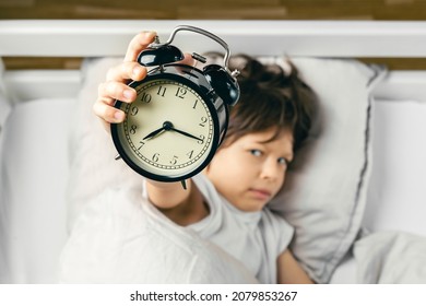 Sleepy Grumpy Boy Is Holding An Alarm Clock That Has Awaken Him Up In The Morning. Focus On The Clock