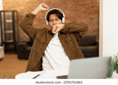 Sleepy Korean Student Guy Using Laptop And Yawning Tired Of Learning Online, Doing Homework Wearing Headphones At Home. Educational Fatigue And Overwork At School Concept - Powered by Shutterstock