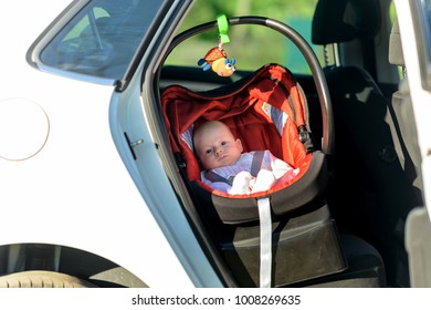 Sleepy Little Baby In A Colorful Red And Black Carrycot On The Back Seat In A Car Viewed Through The Open Door