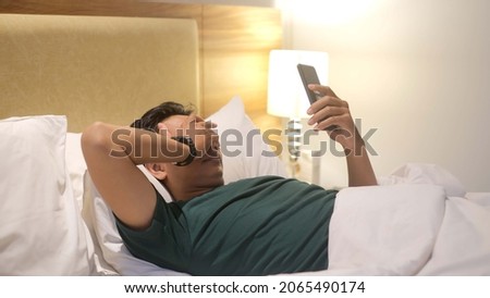 Sleepy man is using smartphone while lying on the bed at night.