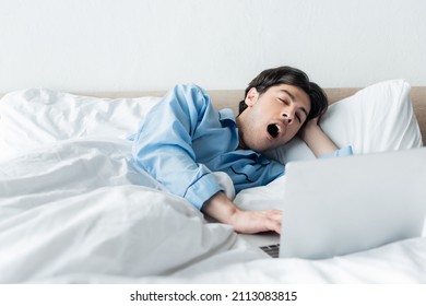 sleepy man yawning near laptop while lying in bed in morning