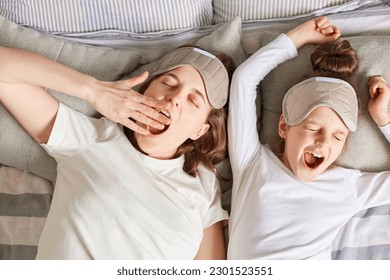 Sleepy mother and daughter sleeping together waking up wearing sleep masks and white shirts yawning lazy morning enjoying happy time together. - Powered by Shutterstock