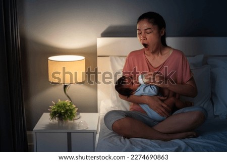 sleepy mother yawning and feeding milk bottle to newborn baby on a bed at night