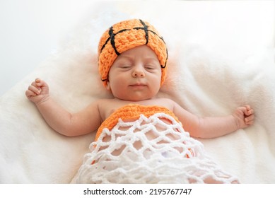 Sleepy Newborn Baby With Blue Eyes Lying On His Back On White Sheet With Orange Hat As A Ball, And Clothes As A Basket Net, Playing Basketball. Top View. Companion Group Game