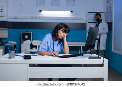 Sleepy Nurse Looking At Computer Screen For Healthcare At Office. Exhausted Woman Working Late At Night With Monitor And Technology For Medical Support. Tired Assistant In Cabinet