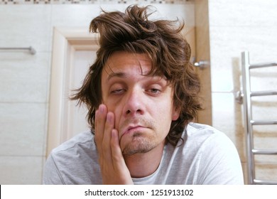 Sleepy shaggy young man looks at the mirror in bathroom in the morning.