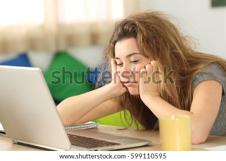 Sleepy student with tousled hair trying to read on line content in a laptop on a desktop in her room in a house interior