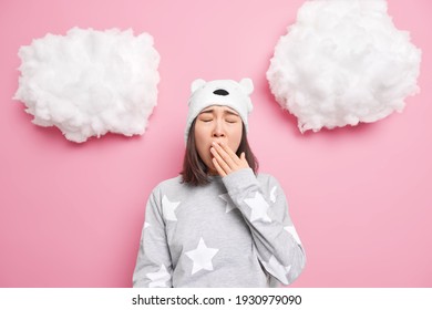 Sleepy Teenage Asian Girl Covers Mouth Yawns Wants To Sleep Has Problem Of Insomnia Dressed In Nightwear Soft Bear Hat Isolated Over Pink Background. Bedtime Energy Charge Concept. Lack Of Vivacity