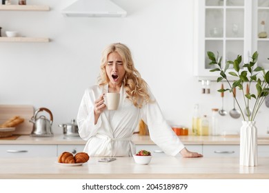 Sleepy tired lazy young pretty caucasian blonde lady in bathrobe wakes up early, drink coffee and yawns in modern kitchen interior, copy space. Lack of sleep, fatigue, bad night dream and breakfast