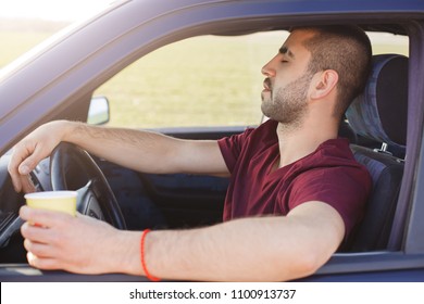 Sleepy unshaven young male sits in automobile, holds cup of coffee, feels tired as drives car with no rest, stops to have break on road, drinks hot beverage. People, tiredness and driving concept