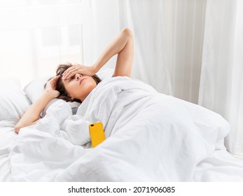 Sleepy woman is lying in bed, completely covered with white blanket. Smartphone used as alarm clock. It's hard to wake up early in morning. Woman does not get enough sleep.