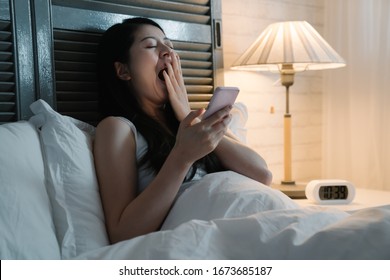 Sleepy Woman Lying On Bed And Yawning But Still Using Smart Phone At Late Night. Conceptual Of Social Media Addiction. Exhausted Asian Chinese Lady Cover Wide Mouth With Hand By Lamp In Dark Bedroom