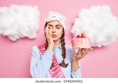 Sleepy woman wakes up very early on her birthday covers face with hand closes eyes holds festive cake wears sleepmask on forehead has two combed pigtails isolated over pink studio background