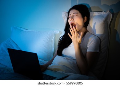 Sleepy Yawn Tired Woman Sitting On Cosy Bed With Laptop In Bedroom. Mixed Race Asian Chinese Model.