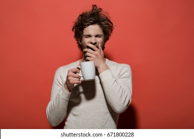 sleepy yawning man with cup of tea or coffee has uncombed hair in underwear on red background, morning refreshment and drink