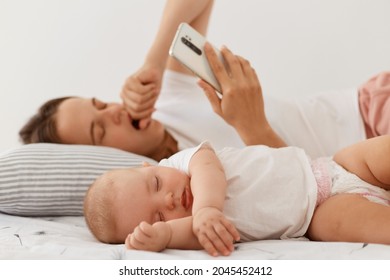 Sleepy Young Adult Female With Dark Hair Lying In Bed With Small Cute Sleeping Baby, Woman Yawning And Covering Mouth With Palm, Using Cell Phone For Checking Emails.