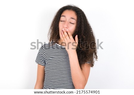 Sleepy young beautiful brunette woman wearing striped t-shirt over white wall yawning with messy hair, feeling tired after sleepless night, yawning, covering mouth with palm.