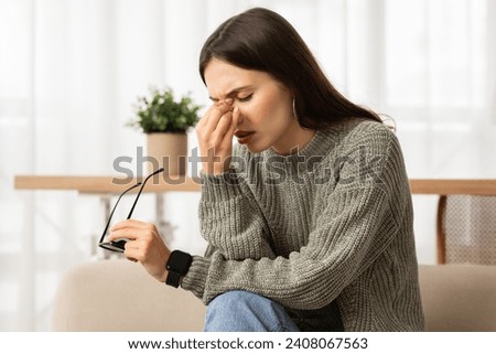 Sleepy young caucasian woman holding glasses rubbing eyes, feels tired after working on computer, sitting on couch at home. Exhausted lady suffering from ocular diseases, eye strain.