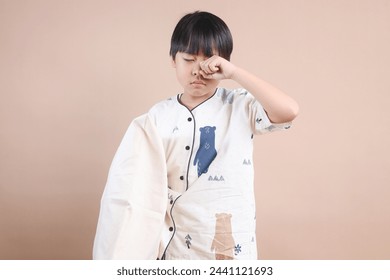 Sleepy Young School Boy Wearing Pajamas Holds Pillow and Rubbing Eye Isolated on Beige Background - Powered by Shutterstock