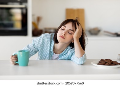 Sleepy young woman drinking coffee, feeling tired, suffering from insomnia and sleeping disorder. Sad female sitting in modern kitchen interior, empty space - Shutterstock ID 2059077560