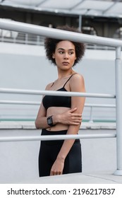 Slender African American Woman In Black Sportswear And Fitness Tracker Looking Away Near Fence