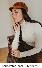 Slender Asian Girl In Corduroy Cap, Brown Pants And Glasses Sits On Chest Of Drawers