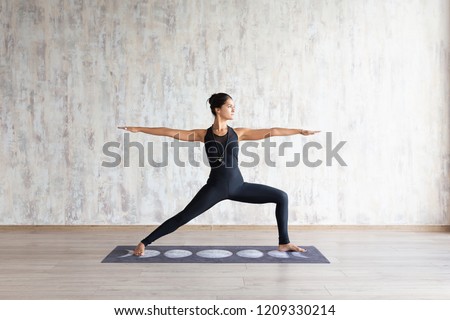 Slender asian girl standing in yoga asana on mat against concrete wall. Peaceful focused girl standing in Warrior Two exercise, Virabhadrasana pose, wearing black sportswear. No stress concept.