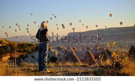 slender attractive young woman at dawn in pink hat, leather jacket and tight bell-bottomed jeans looks at parade of balloons in sky over Turkey. Many balloons with balloonists are navigating in sky.